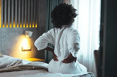 Person sitting on a bed with hands placed on the lower back